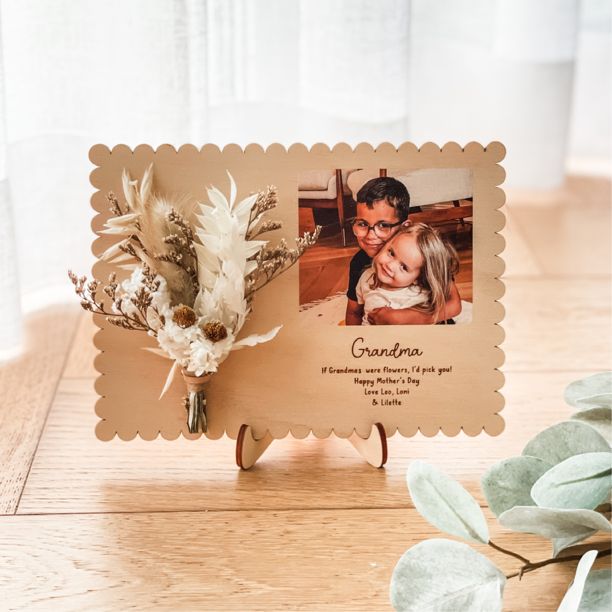 Decorative photo frame with a photo of two children and text, placed on a wooden surface with plants with a happy Mother's Day message