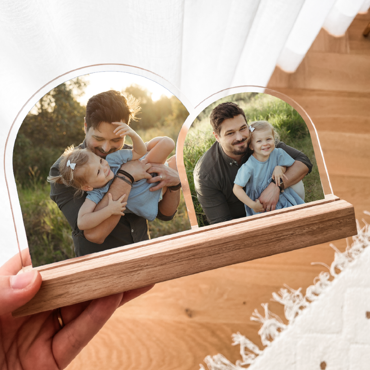 A hand holds a double-arch photo frame with a wooden base, displaying two arched photo slots with clear borders. The photos inside show a man and children in outdoor settings.